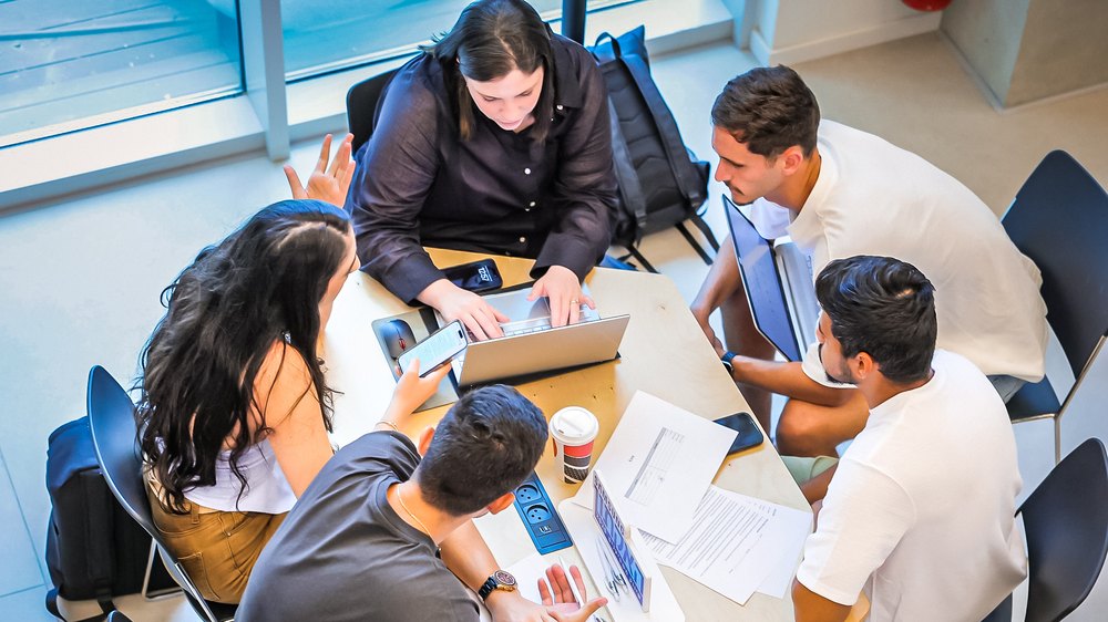 Students working together around a table, laptops and papers spread between them