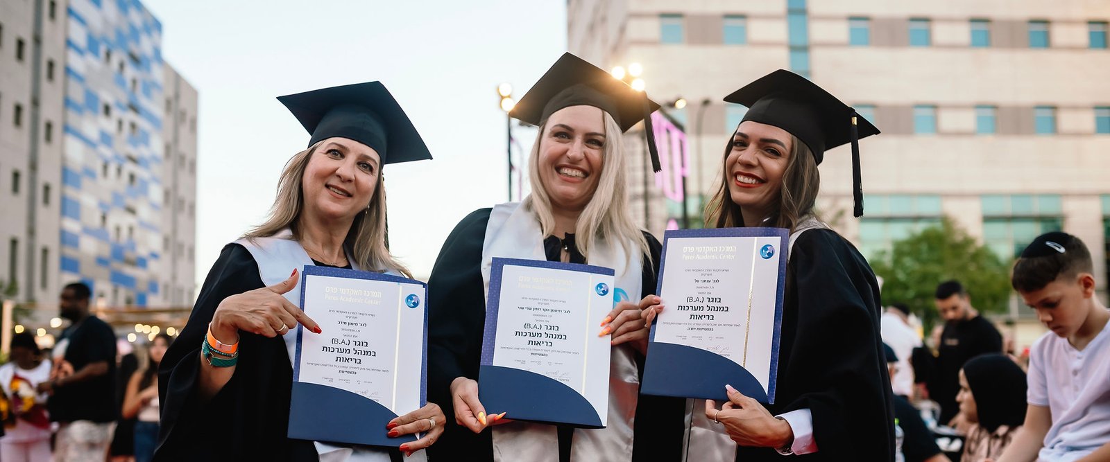 Three women in graduation caps and gowns holding their diplomas at the Peres Academic Center ceremony